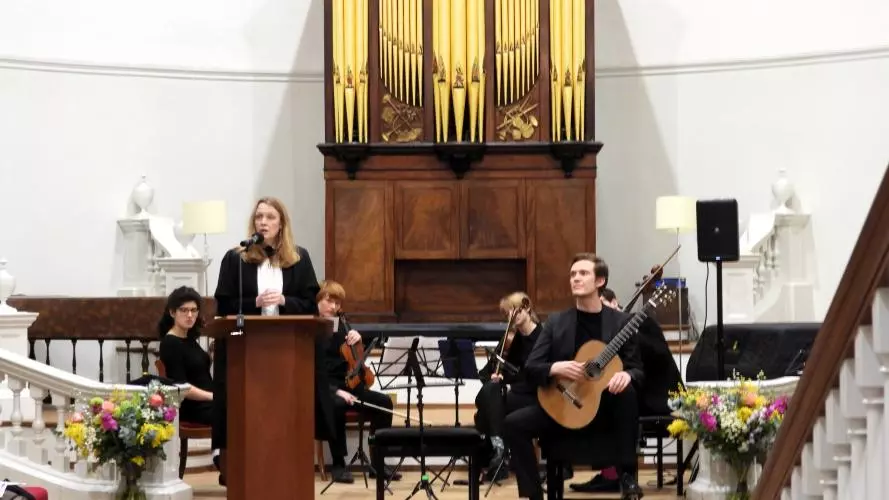 Professor Laura Tunbridge delivering lecture, with student musician on stage behind