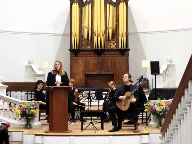 Professor Laura Tunbridge delivering lecture, with student musician on stage behind