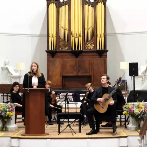 Professor Laura Tunbridge delivering lecture, with student musician on stage behind